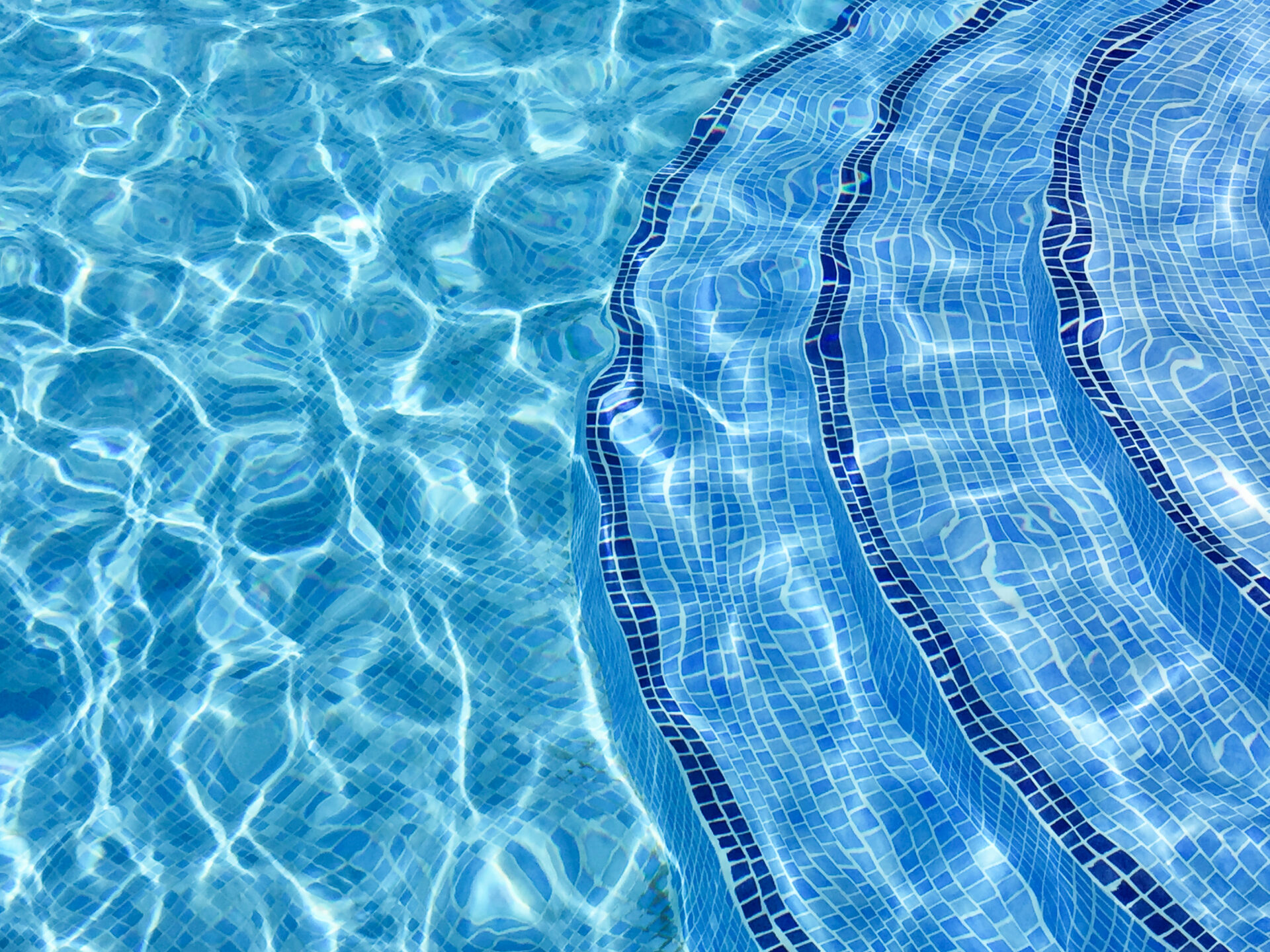 Curved steps into swimming pool, view from above through water on blue mosaic tiles. Distortion. Refraction